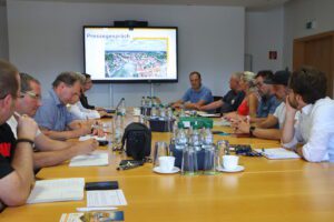 Pressegespräch zum Landeserntedankfest 2025: An einem großen Konferenztisch sitzen etwa zwölf Personen in konzentrierter Gesprächsatmosphäre. Auf dem Tisch stehen zahlreiche Wasserflaschen, Gläser, Kaffeetassen und Notizblöcke. Im Hintergrund zeigt ein großer Bildschirm den Titel „Pressegespräch“ mit einem Luftbild von Hoyerswerda. Die Teilnehmenden wirken engagiert und aufmerksam, es wird mitgeschrieben und diskutiert.