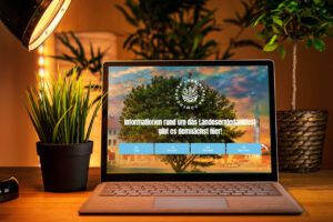 Ein geöffneter Laptop steht auf einem Holztisch, flankiert von zwei grünen Zimmerpflanzen. Auf dem Bildschirm ist die Startseite der Website zum Sächsischen Landeserntedankfest zu sehen. Im Hintergrund ein Baum und Gebäude, darüber der Text: „Informationen rund um das Landeserntedankfest gibt es demnächst hier!“. Darunter ein Countdown mit Tagen, Stunden, Minuten und Sekunden bis zum Website-Start.